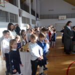 Uczniowie i nauczyciele stoją w sali gimnastycznej podczas spotkania opłatkowego.